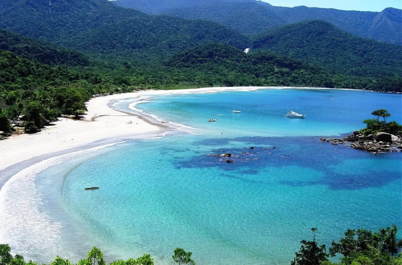 Foto de uma praia paradisíaca em Ilhabela, com águas cristalinas e areia branca, rodeada por montanhas verdes.